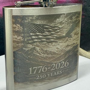 May include: A brushed silver metal flask featuring a laser-engraved design of an American flag, a bald eagle, and mountain peaks. The text 1776-2026 and 250 YEARS is engraved on the front. The flask has a hinged screw-top cap in polished silver.