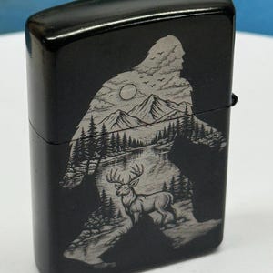 May include: Black metal lighter with a Bigfoot silhouette design. The design features a landscape with mountains, trees, a lake, a deer, and a sun. The lighter is rectangular with rounded edges.