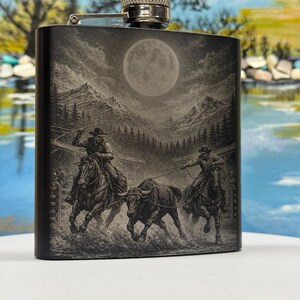 Puede incluir: Una petaca de metal negro mate con una escena grabada con láser de dos vaqueros a caballo lazando un toro en una arena de rodeo. La ilustración detallada incluye una luna llena, picos de montañas y pinos con un tapón de color plateado.