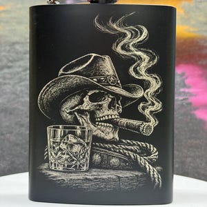 Op de afbeelding: Een matzwarte metalen heupfles met een lasergravure van een schedel met een cowboyhoed die een sigaar rookt, met een glas whisky en een opgerold touw op een houten ondergrond, afgewerkt met een zilveren schroefdop.