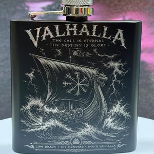 Könnte beinhalten: Ein mattschwarzer Metallflachmann mit silbernem Schraubverschluss. Die Vorderseite zeigt ein eingraviertes Wikingerschiff auf stürmischer See mit einem Vegvisir-Symbol auf dem Segel. Der Text lautet: VALHALLA, THE CALL IS ETERNAL, THE DESTINY IS GLORY, LIVE BRAVE, DIE HONORED, REACH VALHALLA.