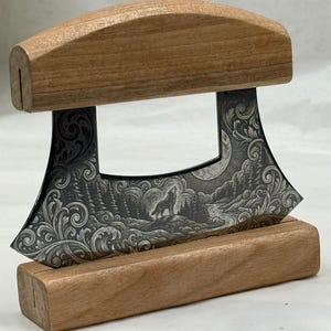 May include: A crescent-shaped mezzaluna knife with a wooden handle and a matching wooden base. The metal blade features a detailed laser-engraved illustration of a wolf howling at a moon, surrounded by a forest landscape and ornate scrollwork.
