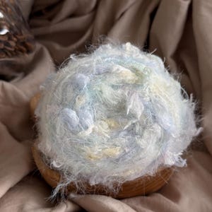 May include: A ball of fluffy yarn in shades of blue, white, and yellow sits in a small wooden bowl. The yarn has a soft, textured appearance, perfect for knitting or other fiber crafts. The background is a neutral, light brown fabric.