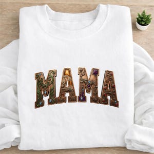 May include: White sweatshirt with the word "MAMA" in a decorative, steampunk-style font. The letters are brown, green, and gold, adorned with jewels and gears. The sweatshirt is folded on a light-colored surface.