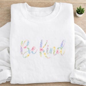May include: A white sweatshirt featuring the phrase "Be Kind" in a floral design. The text is in a cursive font with pastel pink, yellow, blue, and green flowers and leaves. The sweatshirt is folded on a light-colored surface.