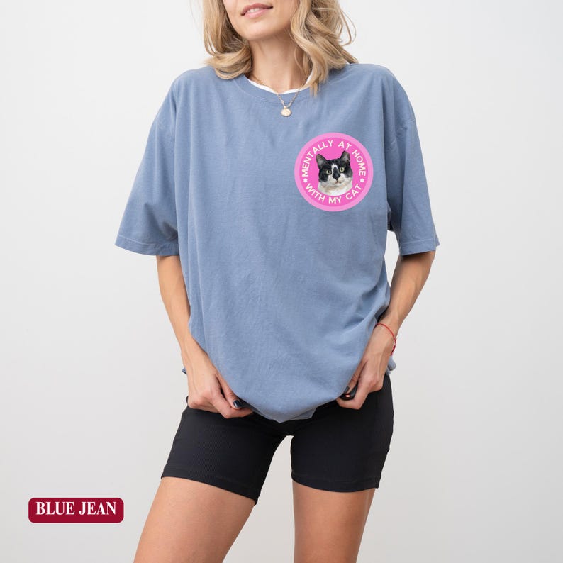 May include: A blue short-sleeved t-shirt with a round graphic of a cat and the text "MENTALLY AT HOME WITH MY CAT". The shirt is paired with black shorts. The text "BLUE JEAN" is at the bottom.