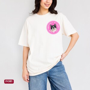 May include: Ivory-colored t-shirt with a round pink graphic featuring a black and white cat and the text "Mentally at home with my cat." The shirt has short sleeves and a relaxed fit.