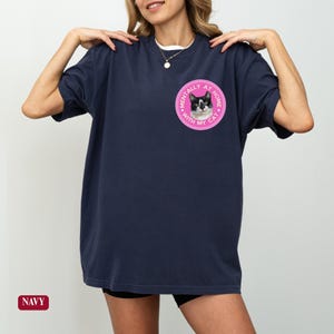 May include: Navy blue t-shirt with a round pink graphic featuring a black and white cat and the text "MENTALLY AT HOME WITH MY CAT". The shirt is a relaxed fit.