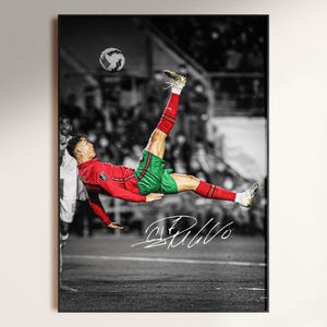 May include: A framed black and white action shot of a soccer player in mid-air, kicking a soccer ball. The player wears a red jersey, green shorts, and white socks. The image includes the player's signature.