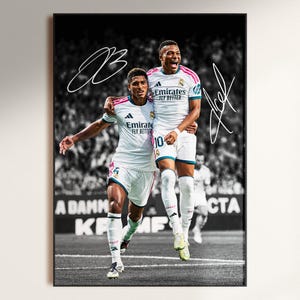 May include: A framed poster featuring two football players in white Real Madrid kits with Emirates Fly Better branding. The players are celebrating on a pitch with a blurred crowd in the background. The image includes two white signatures overlaid on the print.