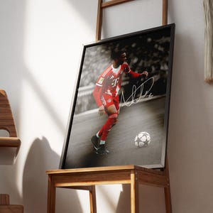 Pode incluir: Fotografia a preto e branco emoldurada de um jogador de futebol com uniforme vermelho, com uma bola de futebol aos seus p&eacute;s. O jogador est&aacute; em a&ccedil;&atilde;o, com uma assinatura branca na imagem. A moldura &eacute; exibida numa mesa de madeira.