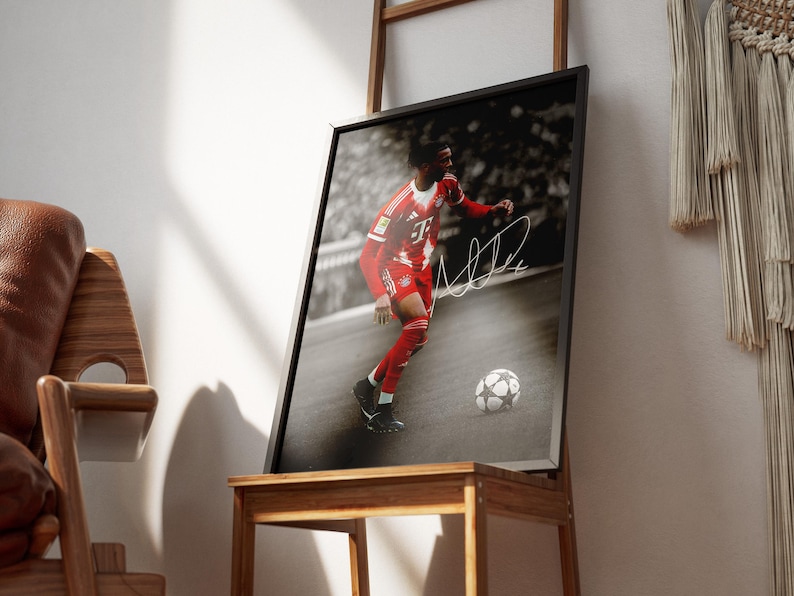Pode incluir: Fotografia a preto e branco emoldurada de um jogador de futebol com uniforme vermelho, com uma bola de futebol aos seus p&eacute;s. O jogador est&aacute; em a&ccedil;&atilde;o, com uma assinatura branca na imagem. A moldura &eacute; exibida numa mesa de madeira.