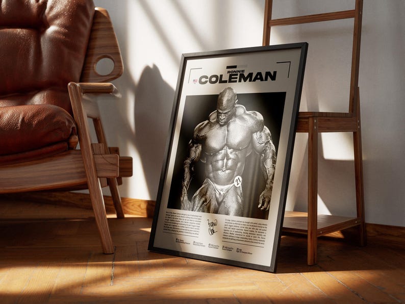 May include: Black framed poster featuring a black and white image of bodybuilder Ronnie Coleman. The poster has the name "RONNIE COLEMAN" at the top and additional text at the bottom. The poster is leaning against a wooden shelf and a brown leather chair.