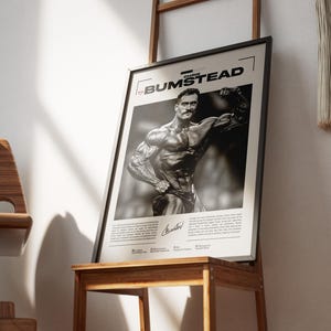 May include: Black and white framed poster featuring bodybuilder Chris Bumstead. The poster is displayed on a wooden stand. The poster includes the name "CHRIS BUMSTEAD" at the top and a signature at the bottom. The image is in a room with a wooden chair and a ladder.