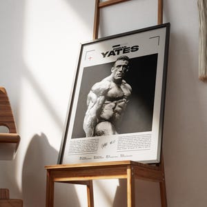 May include: Black and white framed poster featuring a bodybuilder. The poster has the name "Dorian Yates" at the top. The image is of a muscular man. The poster is displayed on a wooden stand.