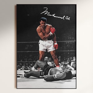 May include: A framed black and white photograph of Muhammad Ali standing over a fallen opponent in a boxing ring. Ali wears white shorts and red boxing gloves. The image features the signature of Muhammad Ali in white script at the top.