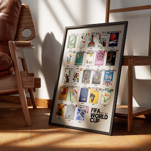 May include: A framed poster featuring FIFA World Cup designs from 1930-2022. The poster has a black frame and is leaning against a wooden stand. The poster is on a wooden floor, with a brown leather chair in the background.