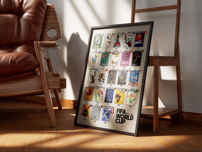 May include: A framed poster featuring FIFA World Cup designs from 1930-2022. The poster has a black frame and is leaning against a wooden stand. The poster is on a wooden floor, with a brown leather chair in the background.