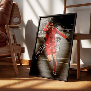 Pode incluir: Impress&atilde;o emoldurada com um jogador de futebol de uniforme vermelho celebrando um gol. A impress&atilde;o tem uma moldura preta e uma assinatura no canto inferior esquerdo. O fundo &eacute; uma imagem em tom s&eacute;pia de um jogo de futebol.