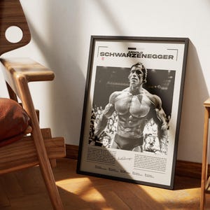 May include: Black framed poster featuring a black and white image of Arnold Schwarzenegger. The poster has the name "ARNOLD SCHWARZENEGGER" at the top. The image shows Schwarzenegger in a bodybuilding pose. The poster is leaning against a wall next to a wooden chair.