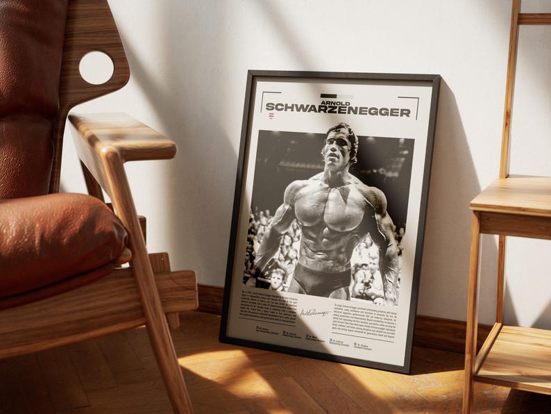 May include: Black framed poster featuring a black and white image of Arnold Schwarzenegger. The poster has the name "ARNOLD SCHWARZENEGGER" at the top. The image shows Schwarzenegger in a bodybuilding pose. The poster is leaning against a wall next to a wooden chair.