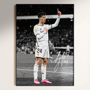 May include: A framed poster featuring a black and white photograph of a soccer player in a white jersey with the number 24, standing on a field with one hand raised. The player wears bright pink cleats and a white signature is visible on the right.