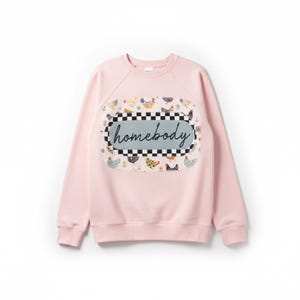 May include: A light pink sweatshirt with a crew neck and long sleeves. The front features a rectangular patch with a black and white checkered border. Inside the border is the word "homebody" in cursive, surrounded by illustrations of chickens.