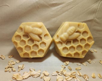 May include: Two hexagonal bars of soap with a honeycomb pattern and a bee design. The soap is light yellow and sits on a beige surface with scattered oats.