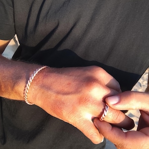 Può includere: Un bracciale color rame metallizzato e un anello abbinato, entrambi caratterizzati da un distinto design a corda intrecciata. Il bracciale è indossato al polso, mentre l'anello è posizionato su un dito, mostrando una consistenza e finitura metallica.