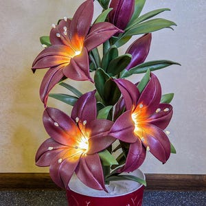 Puede incluir: Lámpara floral decorativa con lirios burdeos con centros iluminados, colocada en una maceta roja con un diseño de mariposa. La lámpara tiene hojas y capullos verdes y un cable de alimentación blanco.