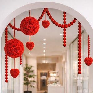May include: An interior hallway with an arched doorway decorated with red rose spheres, heart-shaped ornaments, and strings of red beads. Two white planters with green bushes flank the entrance. The hallway has a gray tiled floor and glass walls.