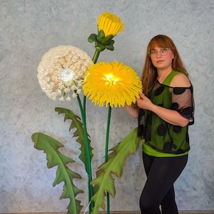 Può includere: Una lampada da terra con tre grandi teste di fiori ispirate ai denti di leone su alti steli verdi. Un fiore è una testa di semi sferica bianca, uno è un fiore giallo brillante e uno è un bocciolo giallo. I fiori hanno grandi foglie verdi.