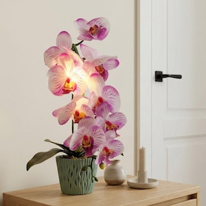 May include: A vibrant orchid plant with pink and purple striped petals in a green textured pot. The plant sits on a wooden nightstand, next to a small vase and a candle.