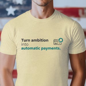 Op de afbeelding: Een lichtgeel t-shirt met de tekst "Turn ambition into automatic payments." in zwart en groen. Het shirt wordt gedragen door een persoon voor een Amerikaanse vlag.