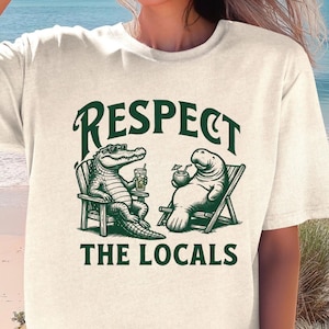 May include: Off-white t-shirt with a green graphic featuring an alligator and a manatee relaxing in beach chairs, holding drinks. The text "RESPECT THE LOCALS" is above and below the illustration. The shirt is a casual, beach-themed design.