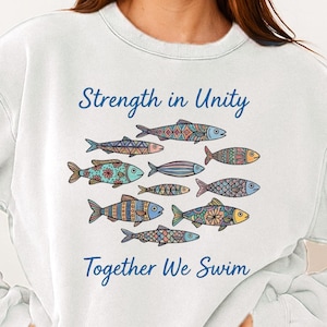 Può includere: Felpa bianca con le scritte "Strength in Unity" e "Together We Swim", sopra e sotto un'illustrazione di pesci colorati e decorati. I pesci sono in tonalità di blu, arancione e verde acqua.