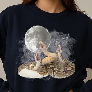 Pode incluir: Camisola azul marinho com um gráfico de três cobras com a boca aberta, uma lua cheia e raios. As cobras são em tons de castanho, bege e amarelo. A camisola é de gola redonda.