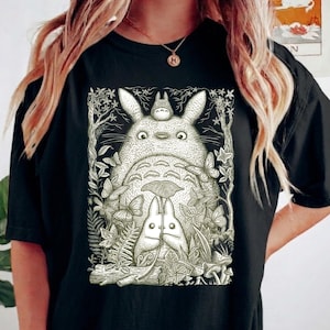 Pode incluir: Camiseta preta com uma ilustra&ccedil;&atilde;o detalhada de Totoro e outros personagens do filme de anima&ccedil;&atilde;o Meu Vizinho Totoro. O gr&aacute;fico &eacute; cercado por folhas, borboletas e outros elementos naturais. O design &eacute; em tons de cinza e branco.