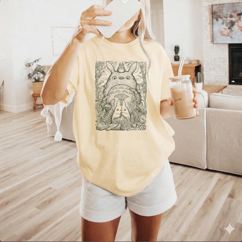 Pode incluir: Camiseta amarelo claro com uma ilustra&ccedil;&atilde;o em preto e branco de Totoro e outros personagens do filme de anima&ccedil;&atilde;o Meu Vizinho Totoro. O gr&aacute;fico est&aacute; centrado na frente da camiseta.