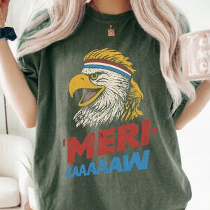 Distressed retro graphic featuring a bald eagle with a squawking open yellow beak and a blonde mullet haircut. The eagle wears a red, white, and blue striped athletic sweatband. Below the illustration, large weathered block text reads "'MERI-" in red, stacked above "CAAAAAW" in bright blue.