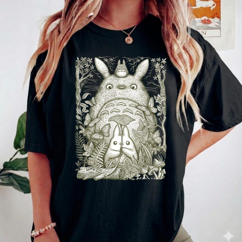 Pode incluir: Camiseta preta com uma ilustra&ccedil;&atilde;o detalhada de Totoro e outros personagens do filme de anima&ccedil;&atilde;o Meu Vizinho Totoro. O gr&aacute;fico &eacute; cercado por folhas, borboletas e outros elementos naturais. O design &eacute; em tons de cinza e branco.