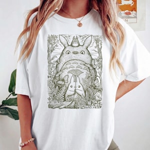 Pode incluir: T-shirt branco com uma ilustra&ccedil;&atilde;o detalhada de Totoro e outras criaturas da floresta. O design &eacute; em tons de verde e preto, com um tema caprichoso inspirado na natureza. O gr&aacute;fico est&aacute; centrado na frente da camiseta.