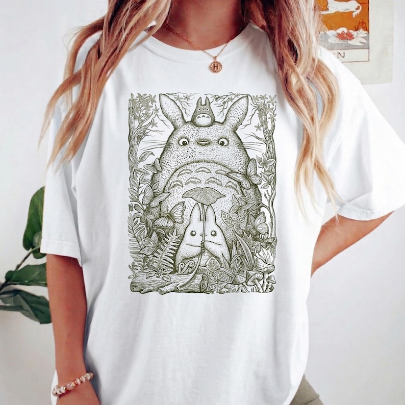 Pode incluir: T-shirt branco com uma ilustra&ccedil;&atilde;o detalhada de Totoro e outras criaturas da floresta. O design &eacute; em tons de verde e preto, com um tema caprichoso inspirado na natureza. O gr&aacute;fico est&aacute; centrado na frente da camiseta.