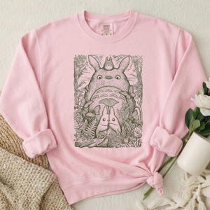Pode incluir: Um moletom rosa claro com uma ilustra&ccedil;&atilde;o em preto e branco de uma cena de floresta com tr&ecirc;s personagens Totoro. O moletom tem mangas compridas e gola redonda. As mangas est&atilde;o enroladas.