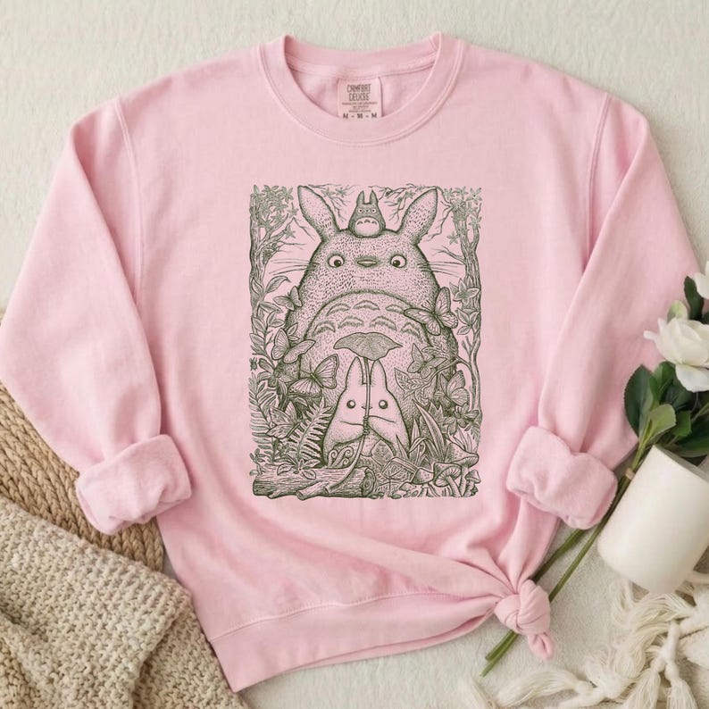 Pode incluir: Um moletom rosa claro com uma ilustra&ccedil;&atilde;o em preto e branco de uma cena de floresta com tr&ecirc;s personagens Totoro. O moletom tem mangas compridas e gola redonda. As mangas est&atilde;o enroladas.