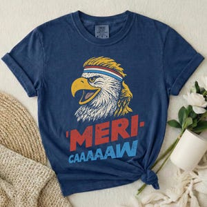 Distressed retro graphic featuring a bald eagle with a squawking open yellow beak and a blonde mullet haircut. The eagle wears a red, white, and blue striped athletic sweatband. Below the illustration, large weathered block text reads "'MERI-" in red, stacked above "CAAAAAW" in bright blue.