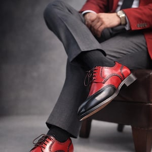 May include: Red and black leather dress shoes with brogue detailing. The shoes have a classic wingtip design and are laced up with brown laces. The shoes are worn with grey trousers and a red blazer.