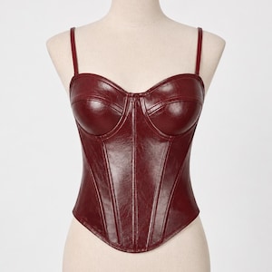 K&ouml;nnte beinhalten: Ein weinrotes Bustier-Top mit d&uuml;nnen Tr&auml;gern. Das Oberteil hat ein strukturiertes Design mit vertikalen N&auml;hten und einem Sweetheart-Ausschnitt. Das Top wird auf einer Schaufensterpuppe pr&auml;sentiert.