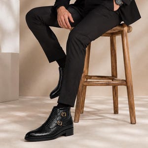 Könnte beinhalten: Schwarze Stiefeletten mit strukturiertem Schlangenledermuster, ausgestattet mit zwei goldfarbenen seitlichen Schnallen und einem niedrigen Blockabsatz. Die Stiefel werden mit einer schwarzen Hose und schwarzen Socken getragen und haben eine spitze Form.