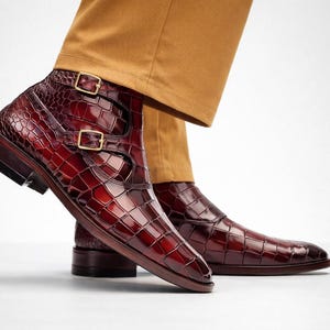 May include: Burgundy leather ankle boots with a crocodile texture and double buckle fastening. The boots have a polished finish and a dark brown sole. The footwear is paired with tan trousers.